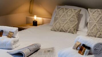 Una cama bien hecha con toallas enrolladas, un periódico y una pequeña botella de loción a un lado.