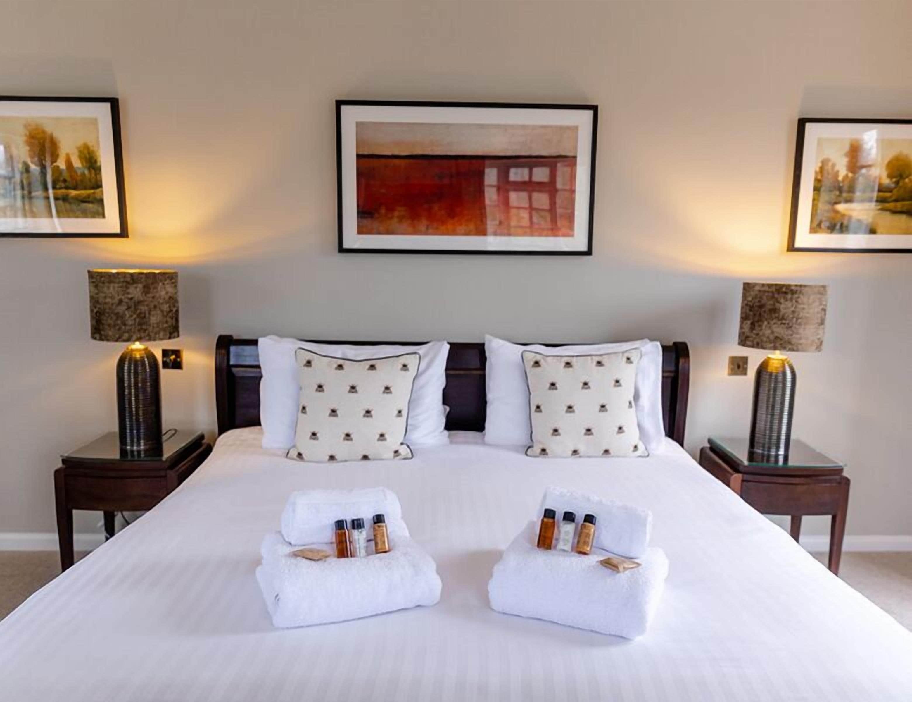 A neatly made bed with two towels folded at the foot, a small table with three bottles, and two lamps on either side.