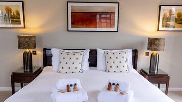 A neatly made bed with two towels folded at the foot, a small table with three bottles, and two lamps on either side.