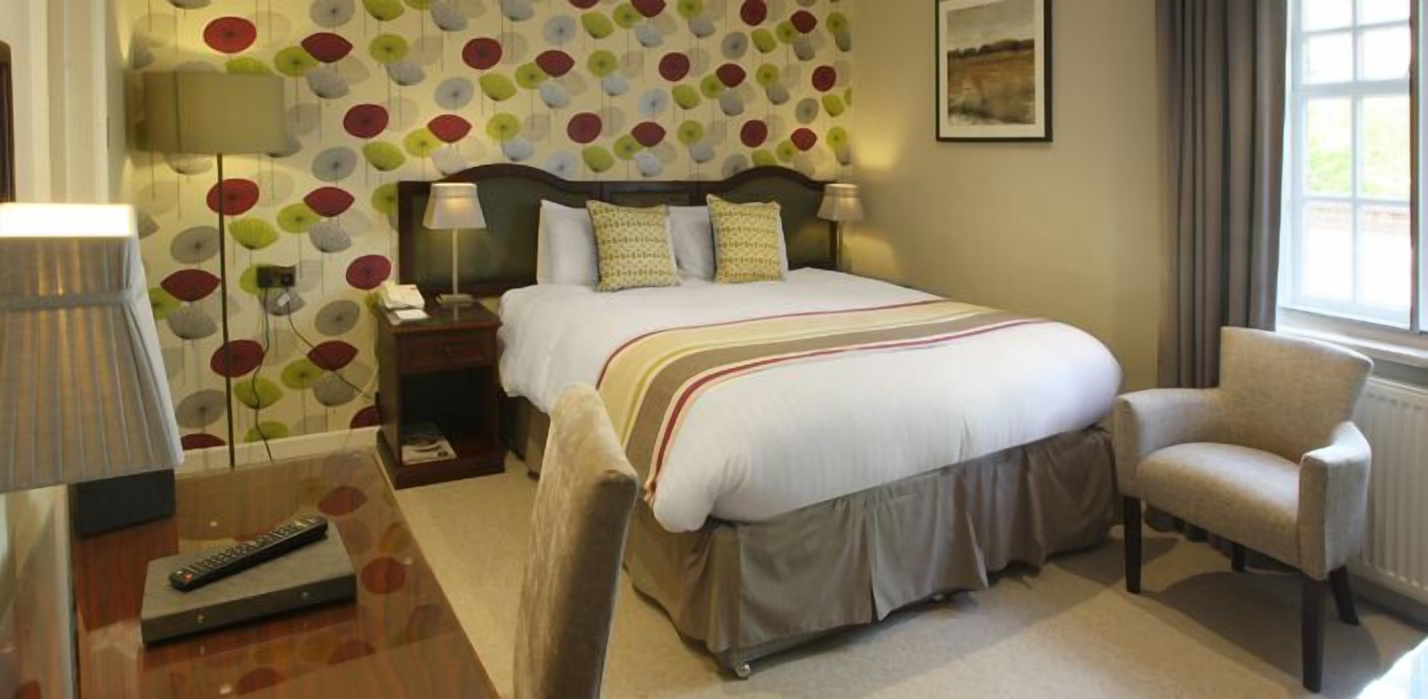 A hotel room with a bed, a chair, a nightstand, a lamp, and a wall with a polka dot pattern.