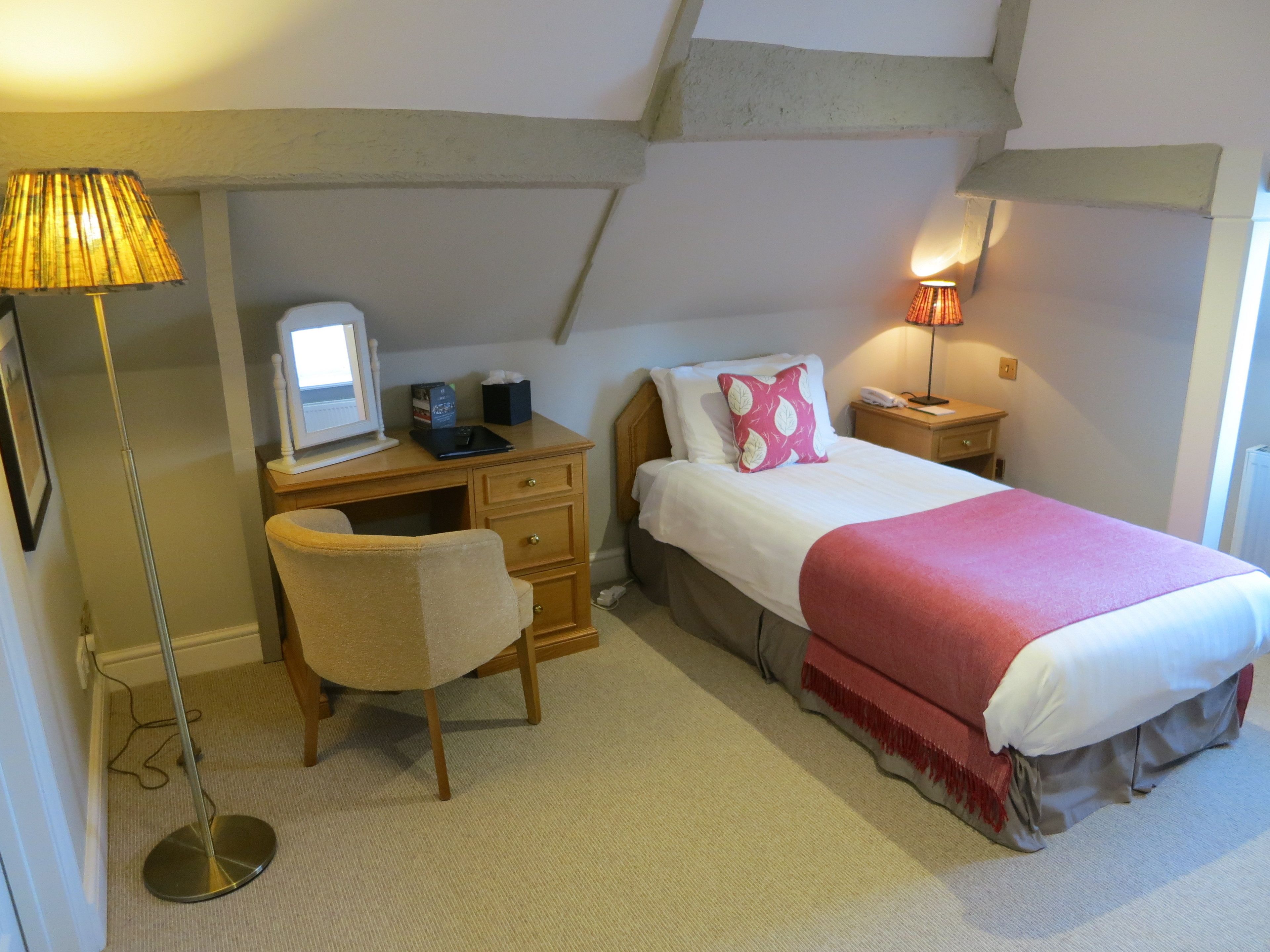 A bedroom with a desk, chair, bed, bedside table, lamp, and a window.