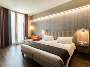 A hotel room with a large bed, a small armchair, a bedside table with a lamp, and a view of the outdoors through the window. - Mercure Firenze Centro (Florence)