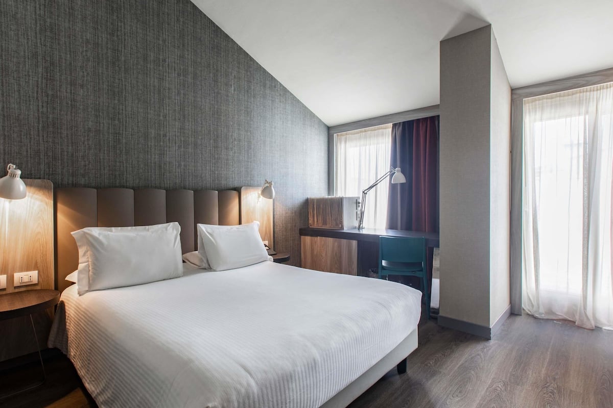 Una habitación de hotel moderna con una cama grande, dos mesitas de noche con lámparas, un escritorio con silla y un amplio ventanal con cortinas.
