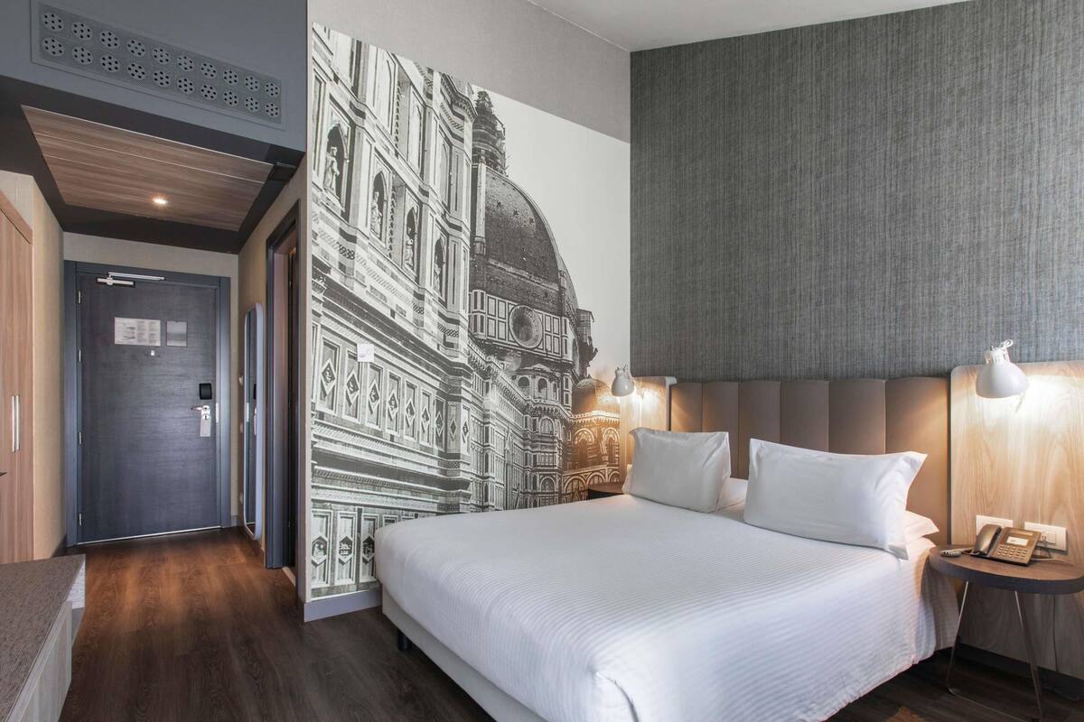 Una habitación de hotel con una cama grande, una mesita de noche con lámpara, un teléfono en la pared y una puerta con picaporte.