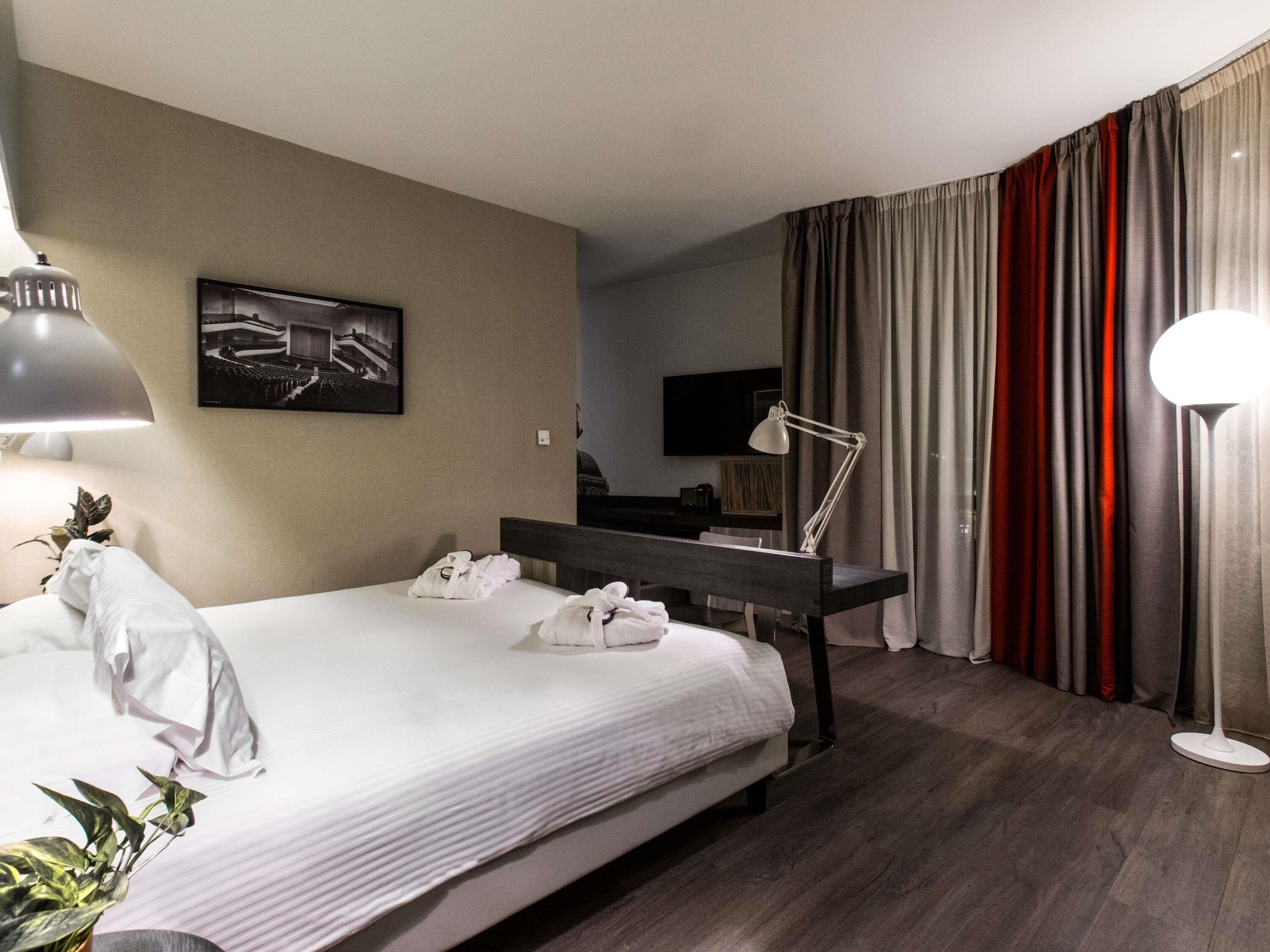 A hotel room with a bed, bedside lamp, a desk, a chair, a TV, and a plant.