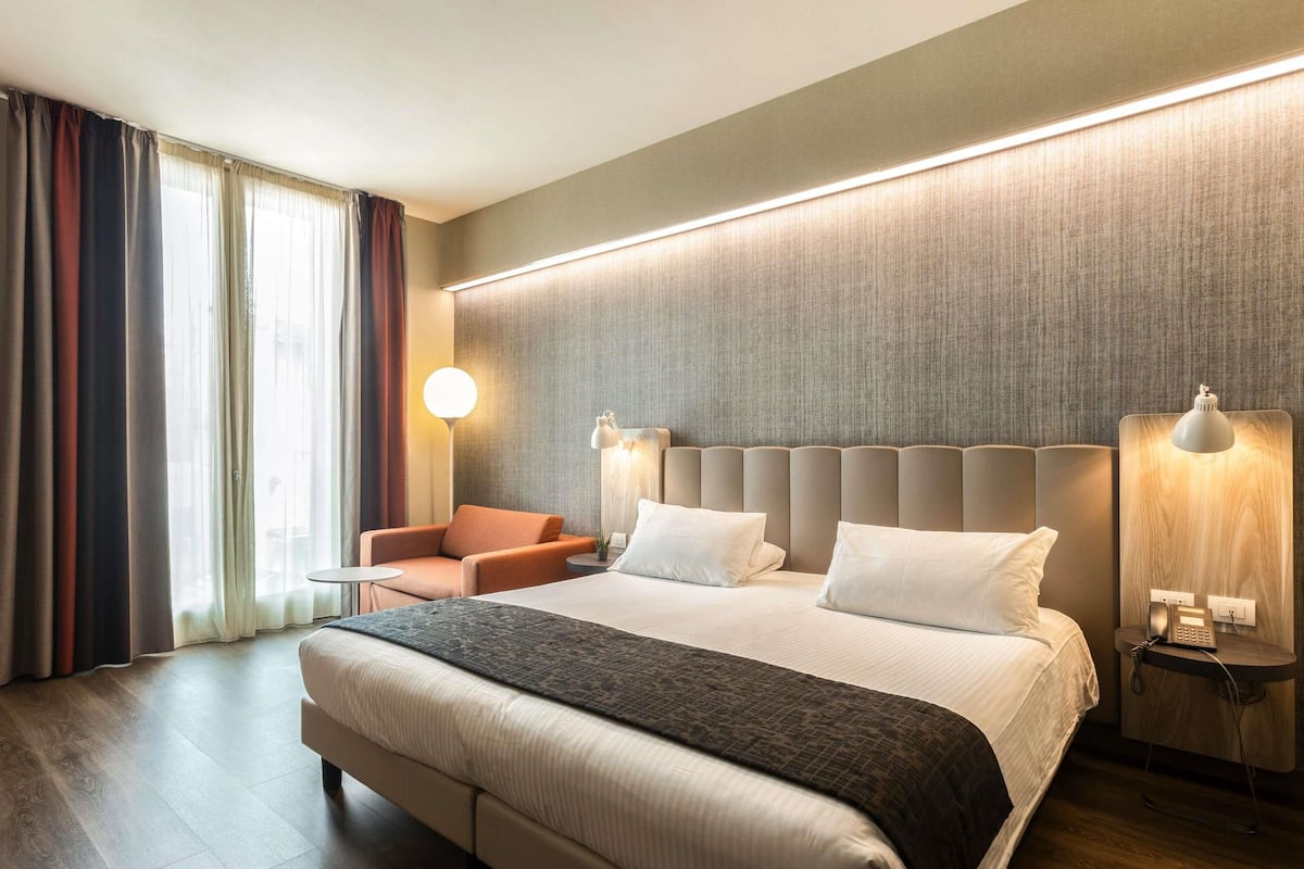 Un cuarto de hotel con una cama grande, un sillón pequeño, una mesita de noche con una lámpara y vistas al exterior por la ventana.