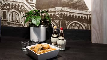 Una mesa de comedor con un tazón blanco de comida frita, dos botellas pequeñas de salsa, un vaso y una planta en maceta.