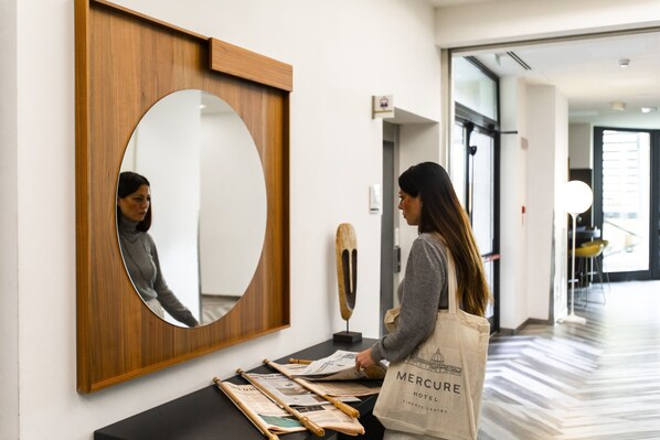 Una mujer de pie en un pasillo con un espejo de marco de madera, una escultura de madera y periódicos sobre una mesa negra.