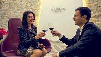 A man and a woman sitting in a leather chair, holding wine glasses, with a stone wall and a sign that reads 'Mercure Hotels' in the background.