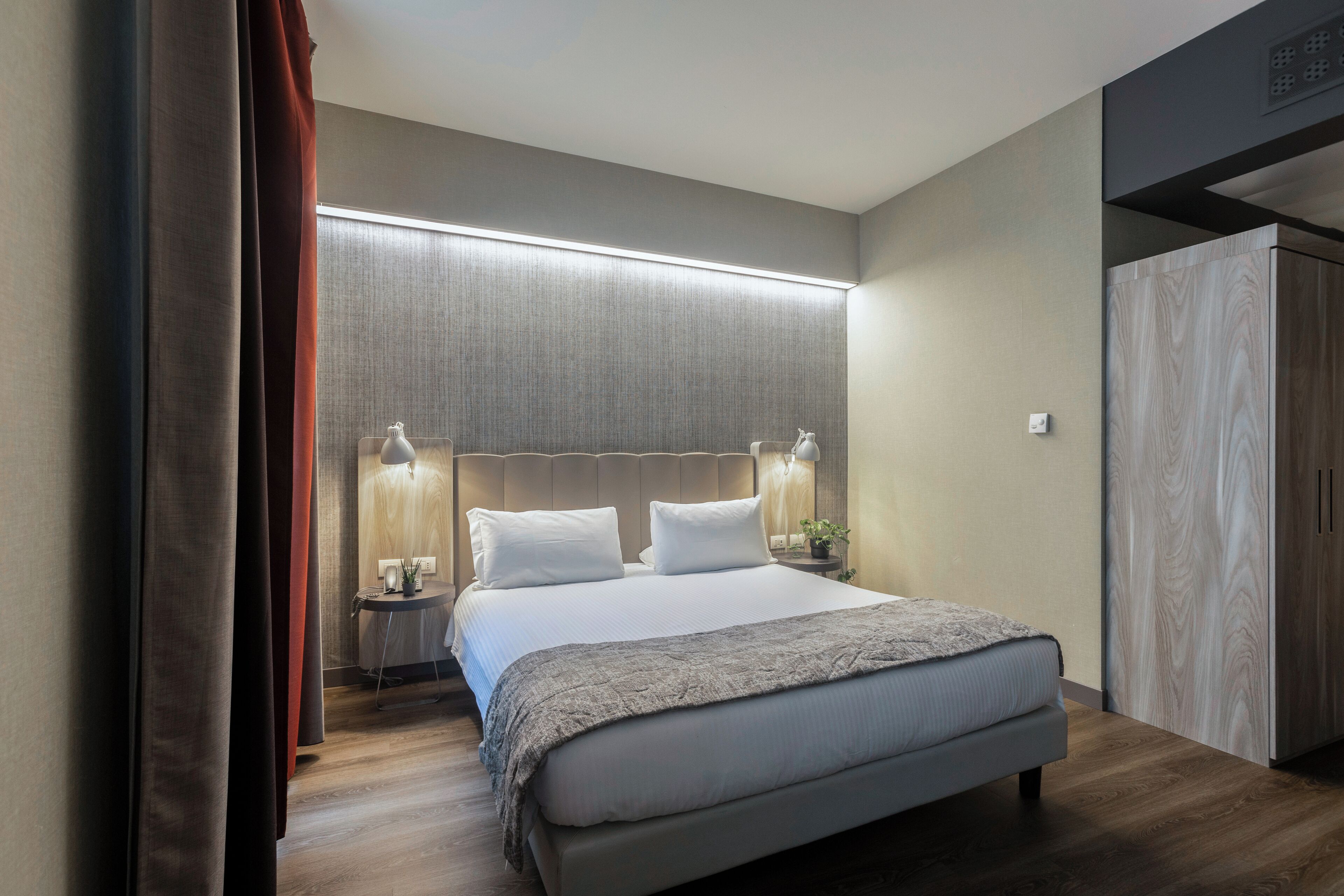 A modern hotel room with a large bed, bedside lamps, a wardrobe, and a bedside table with a lamp.