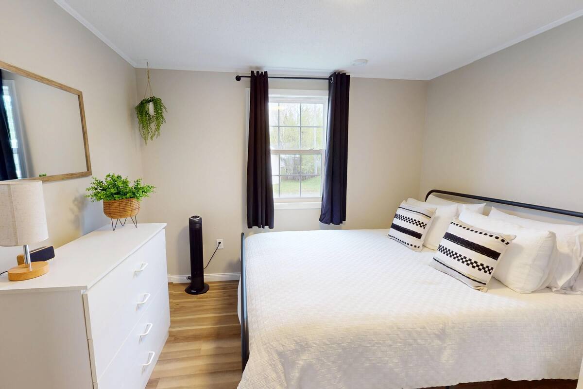 A bedroom with a bed, bedside tables, a dresser, a mirror, a window with curtains, and a hanging plant.