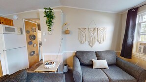 Un espace de vie compact comprenant un coin cuisine, un canapé gris, des décorations murales en macramé et une petite table avec un panier.
