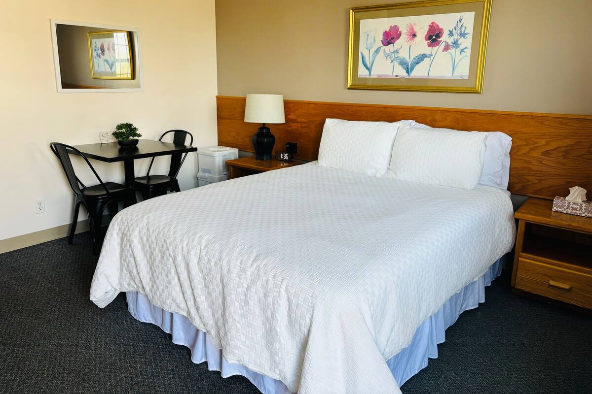 A hotel room with a large bed, a desk with a chair, a nightstand, a lamp, a framed floral artwork, and a small potted plant.