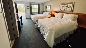 Une chambre d’hôtel avec deux lits, un bureau et une chaise. Il y a une fenêtre avec des rideaux, un tableau au mur et un luminaire au plafond.