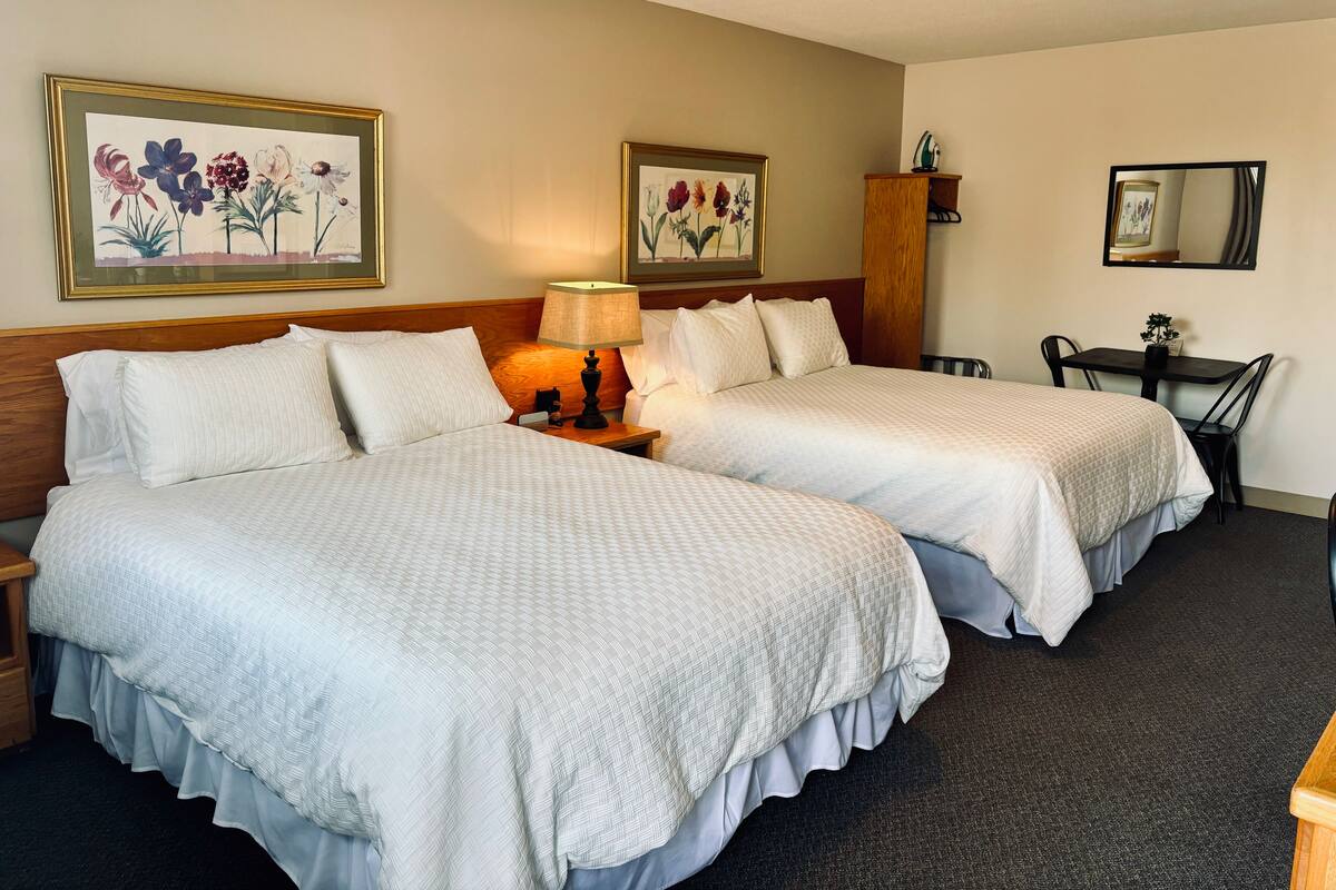 A hotel room with two beds, a nightstand with a lamp, a wooden headboard, a small table with a chair, and two framed floral paintings on the wall.