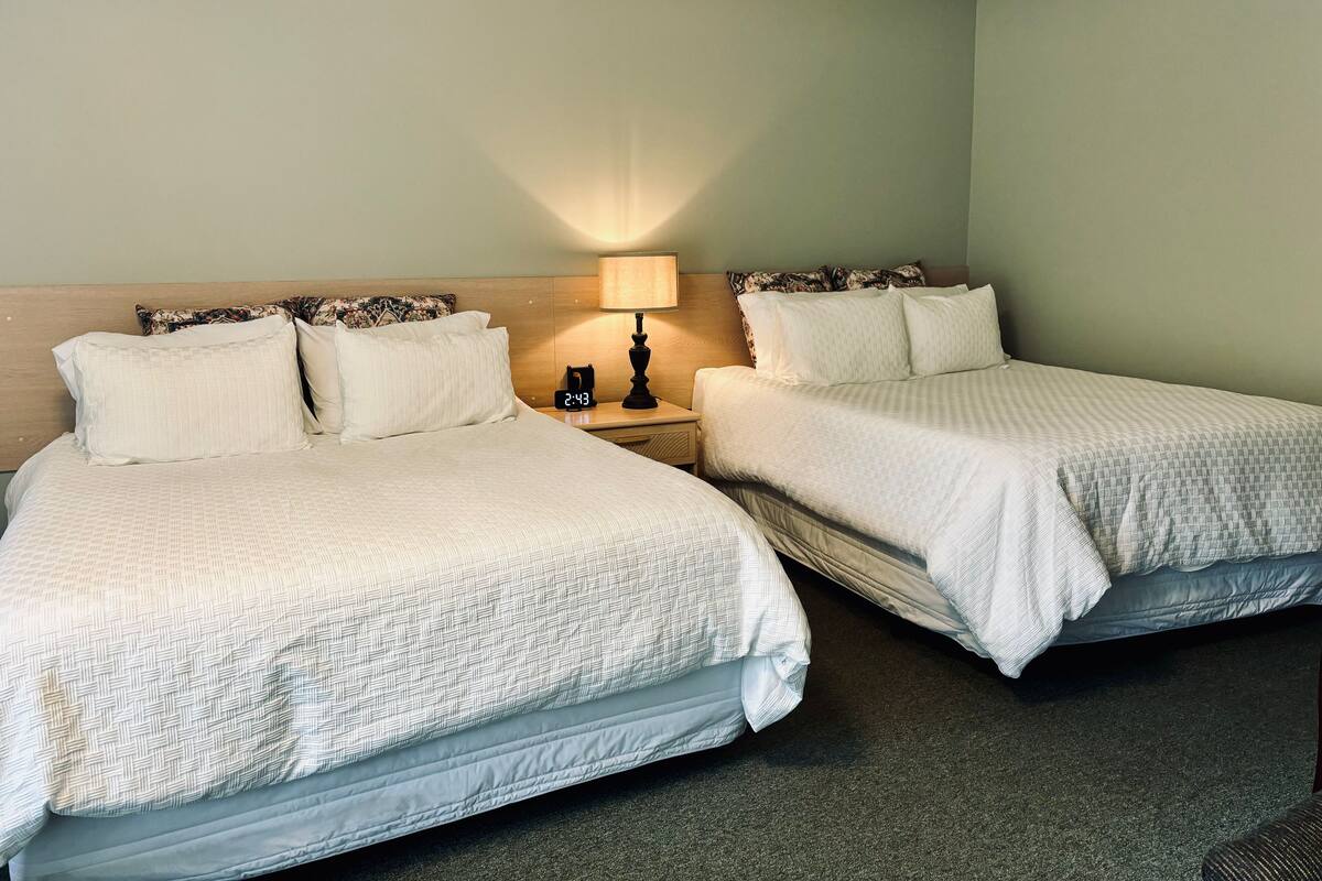 A hotel room with two beds, a nightstand with a lamp, and a clock displaying 2:43.