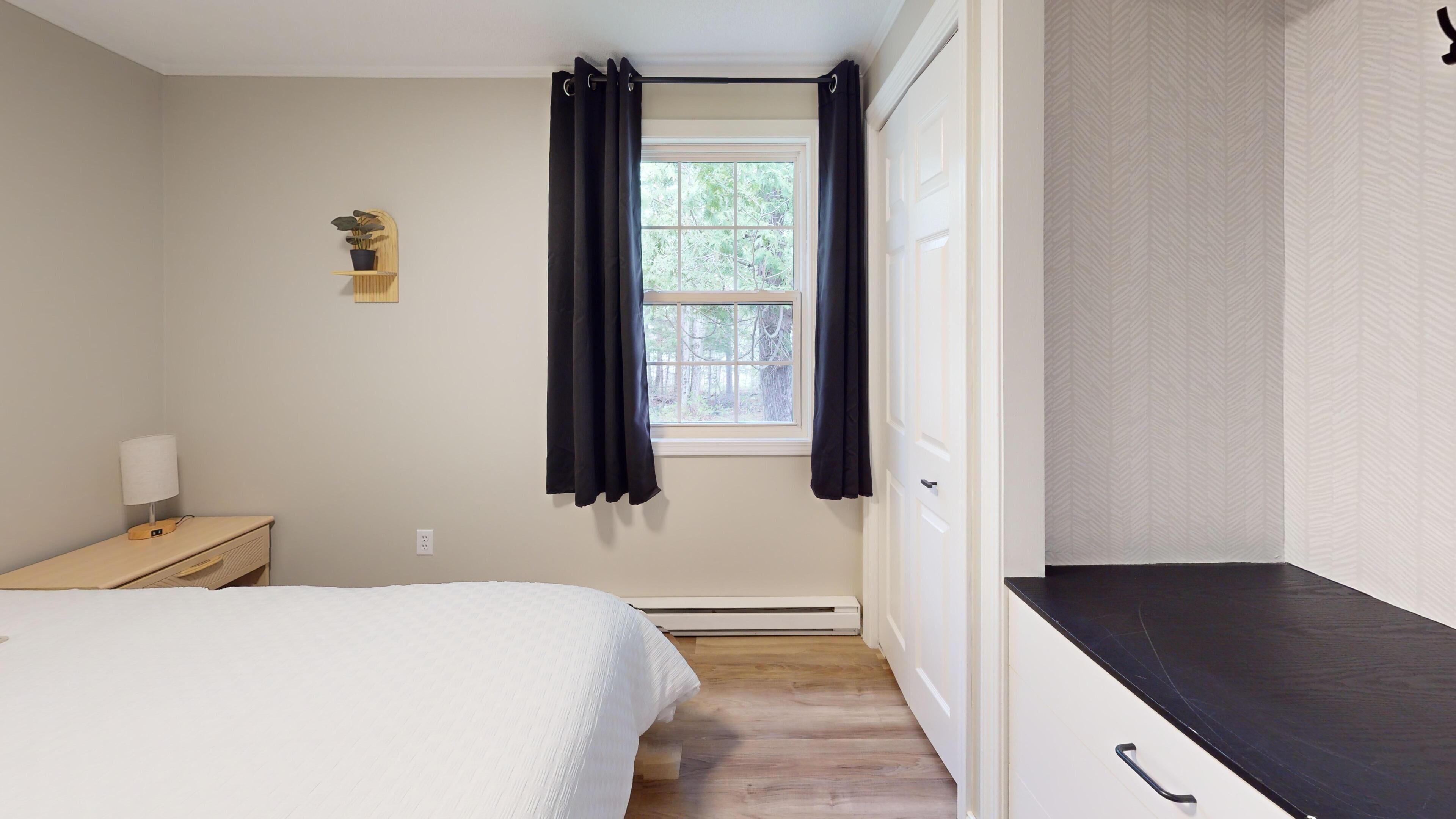A bedroom with a bed, a bedside table with a lamp, a window with curtains, a heater, and a wardrobe.
