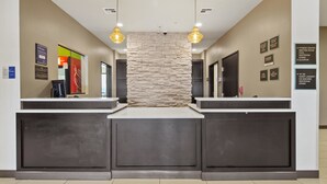 Una zona de recepción con un muro decorativo de piedra, dos mostradores de recepción y señales indicativas para servicios como el gimnasio, la sala de reuniones, la piscina y los baños.