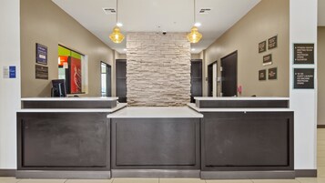 Uma área de recepção com parede decorativa em pedra, dois balcões de recepção e placas indicativas para serviços como centro de ginástica, sala de reuniões, piscina e sanitários.