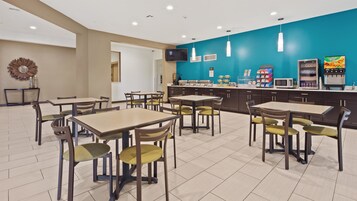 Uma área de refeições moderna com mesas, cadeiras, um balcão com alimentos e uma tela de televisão.