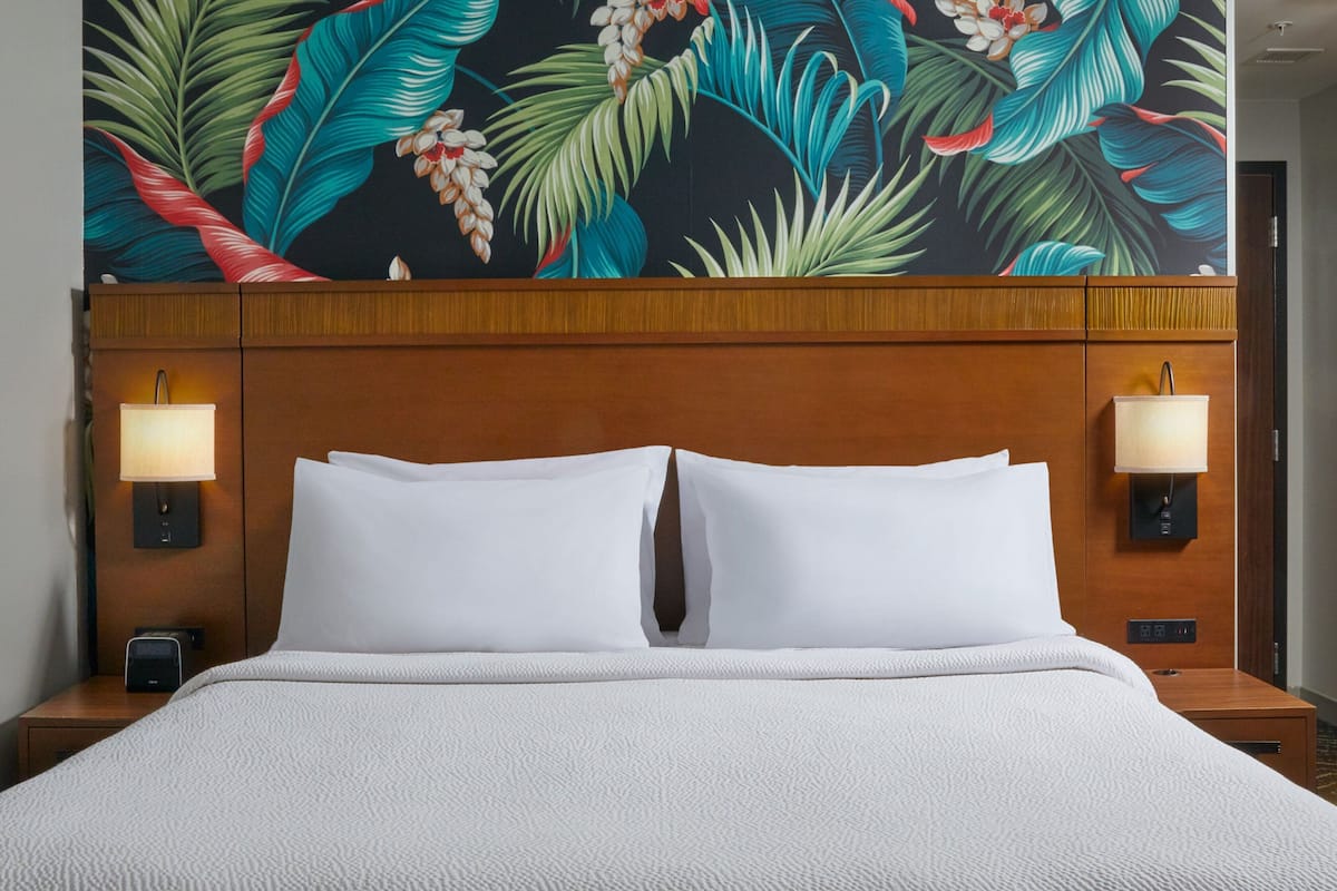 Une chambre d’hôtel avec un grand lit, deux lampes de chevet, une table de nuit et une fresque murale sur le thème des tropiques.