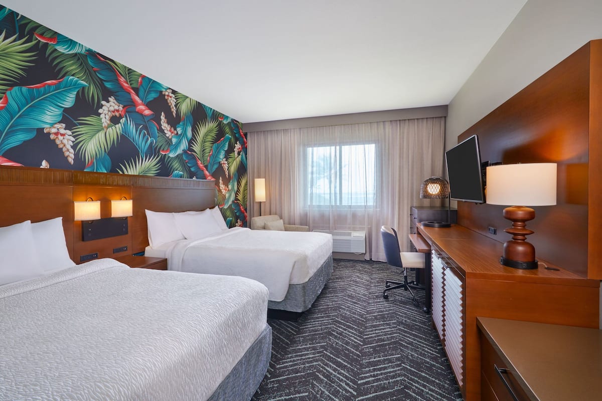 Une chambre d’hôtel avec deux lits, une grande fresque murale aux motifs tropicaux, un bureau avec une lampe et un téléviseur.