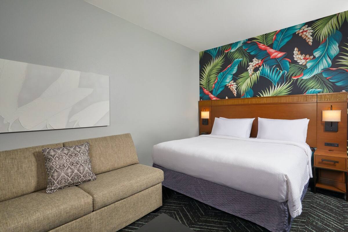 Une chambre d’hôtel avec un lit, un canapé, une table de chevet et un mur orné d’un motif de feuilles tropicales.