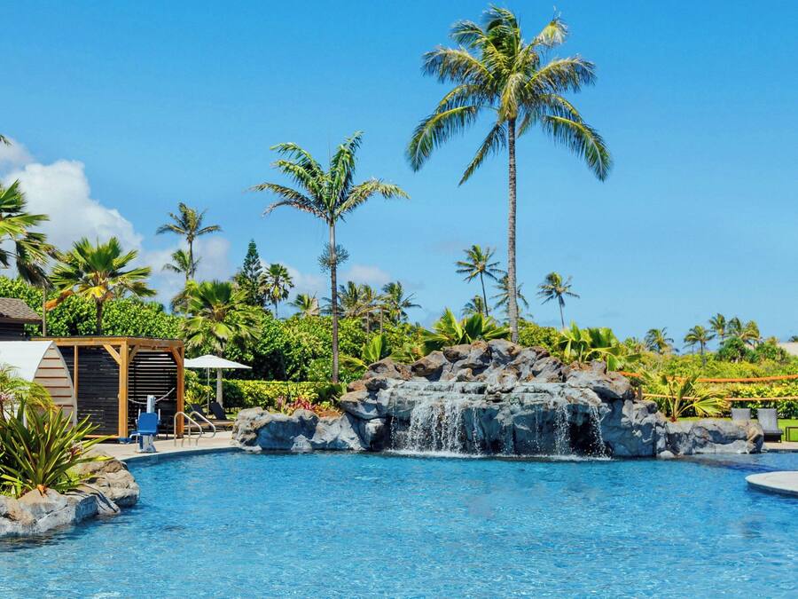 A pool with a waterfall, surrounded by palm trees and a wooden structure.