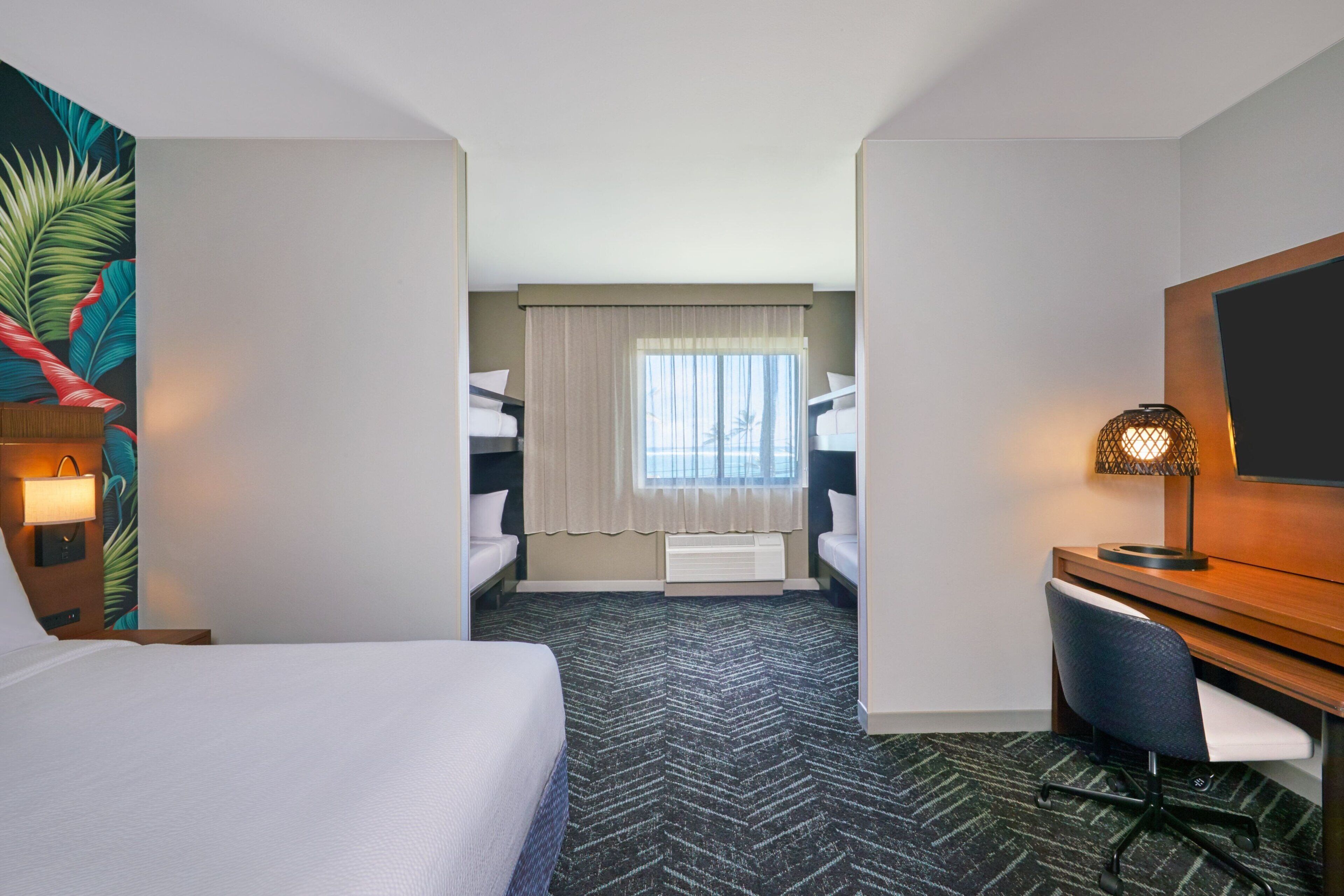 A hotel room with a bed, a desk with a chair, a television, and a window with curtains.