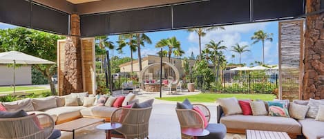 A spacious outdoor seating area with wicker furniture and a stone pillar, overlooking a tropical landscape with palm trees and a pool.