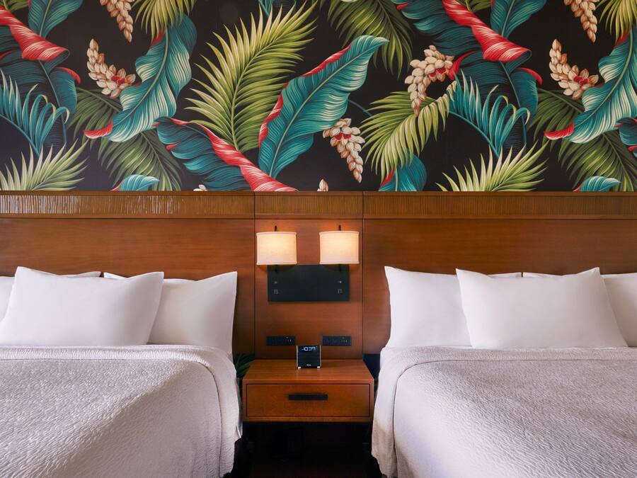 A hotel room with two beds, a nightstand with a clock, and a wall with a tropical leaf and flower mural.