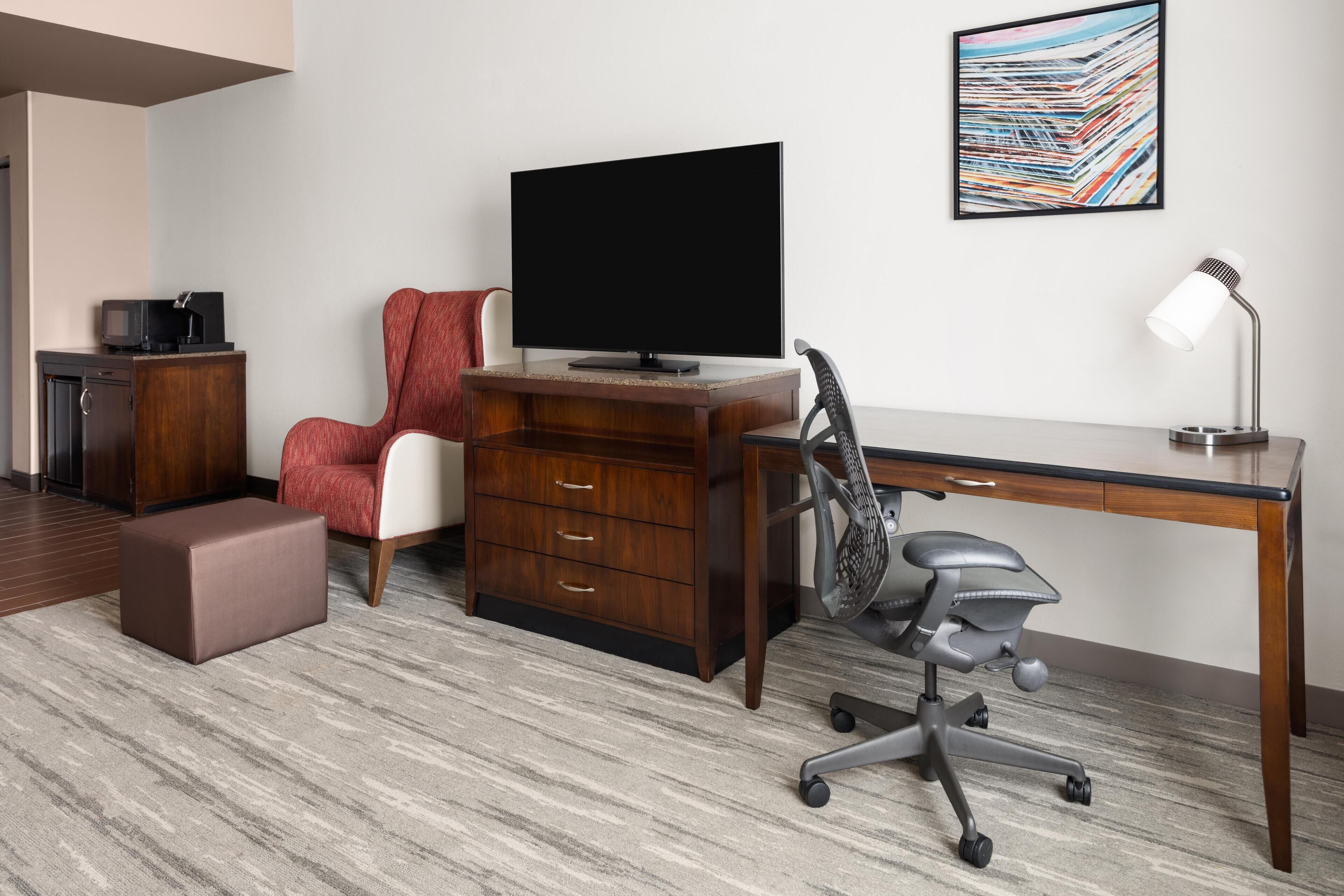 Un espacio de oficina moderno con un escritorio de madera, una silla roja, una silla de oficina negra, un televisor, una lámpara de escritorio y un archivador.