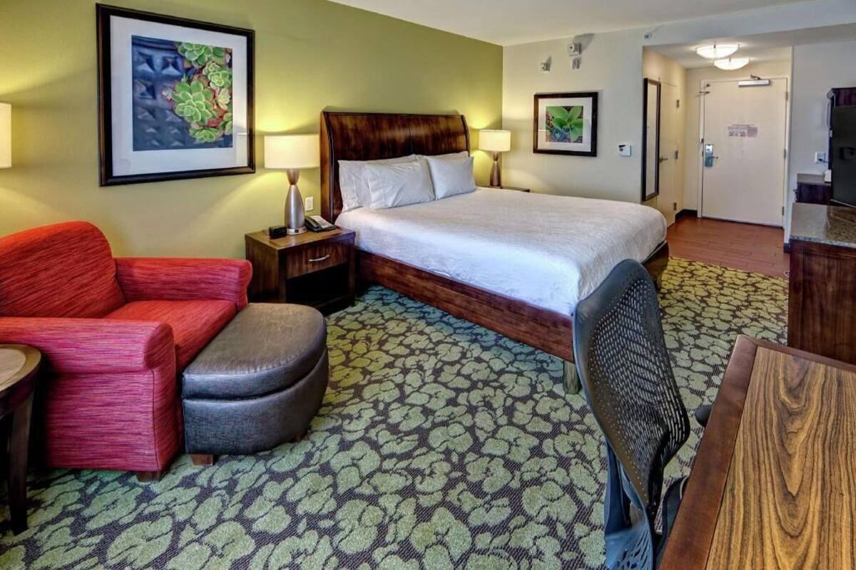A hotel room with a bed, a red sofa, a grey ottoman, a wooden table, and a patterned carpet.
