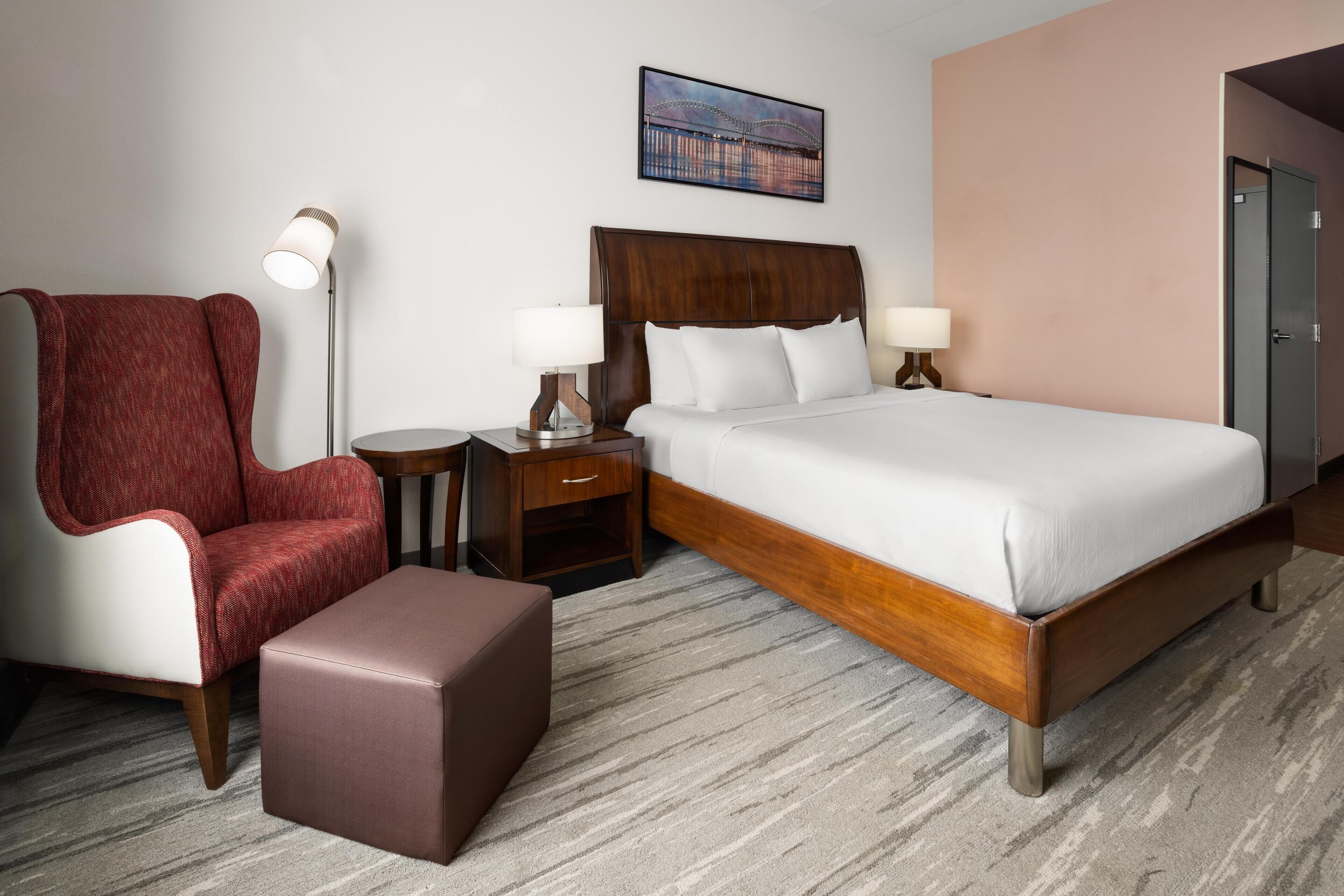 Een hotelkamer met een groot bed, een rode fauteuil, een klein tafeltje en een lamp.