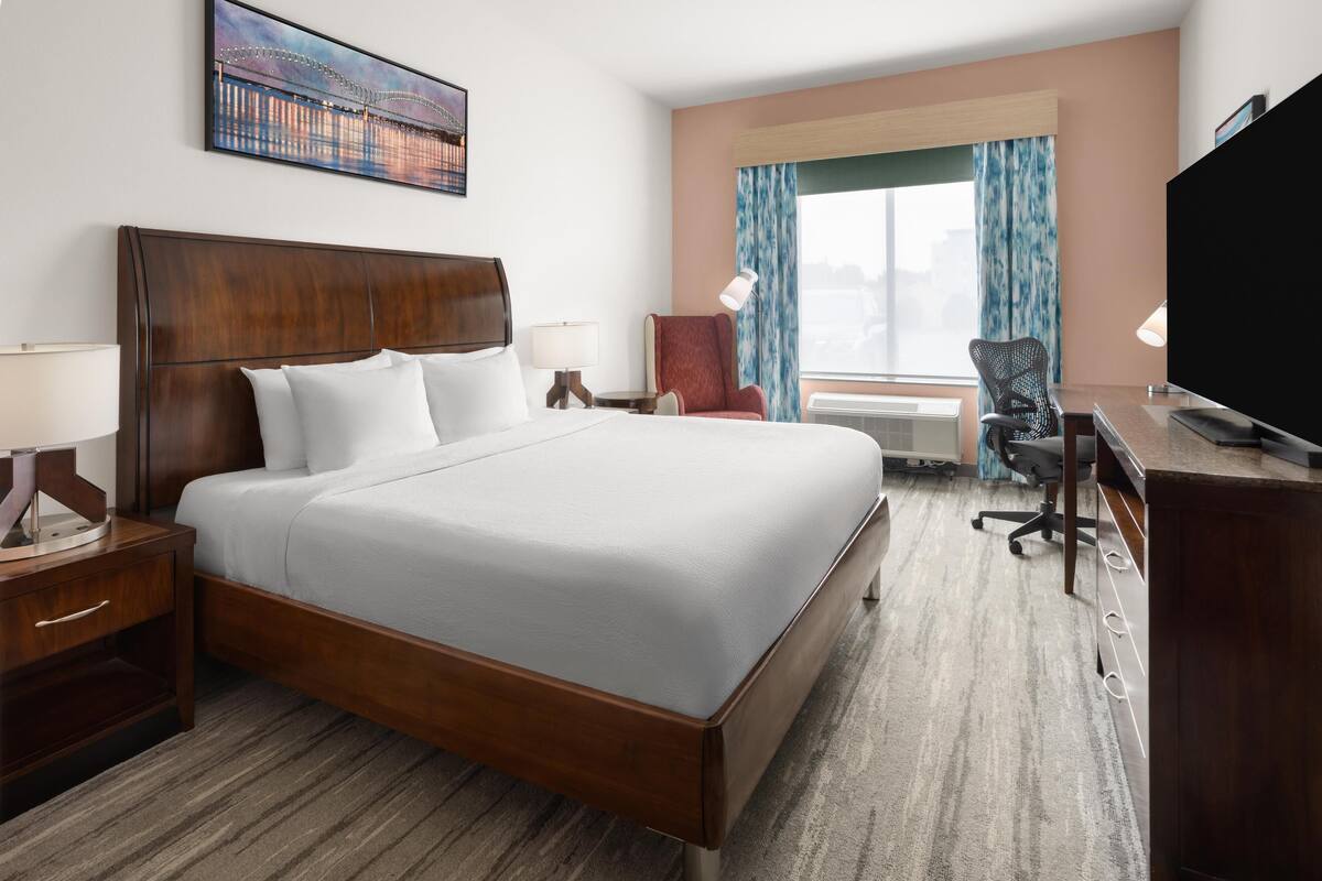 A hotel room with a large bed, a desk, a chair, a TV, and a window with curtains.