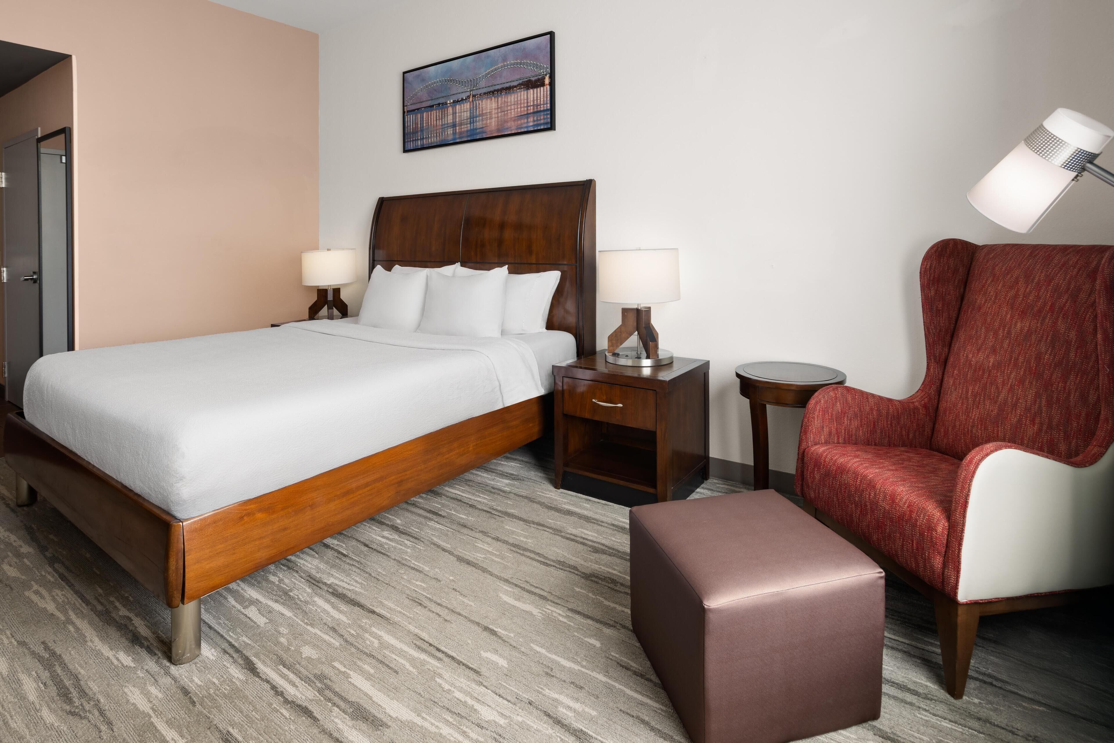 A hotel room with a wooden bed, two bedside tables with lamps, a red armchair, and a small ottoman.