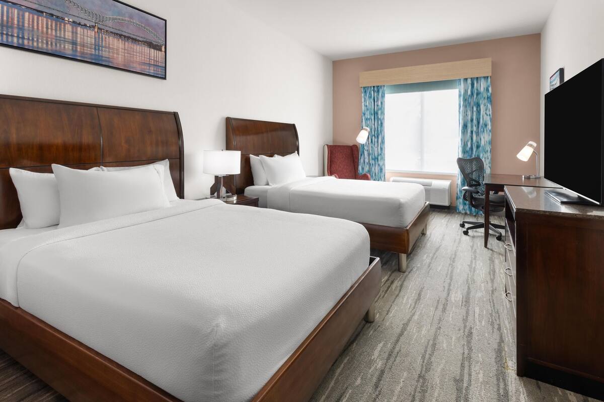 A hotel room with two beds, a desk, a chair, a TV, and a window with curtains.