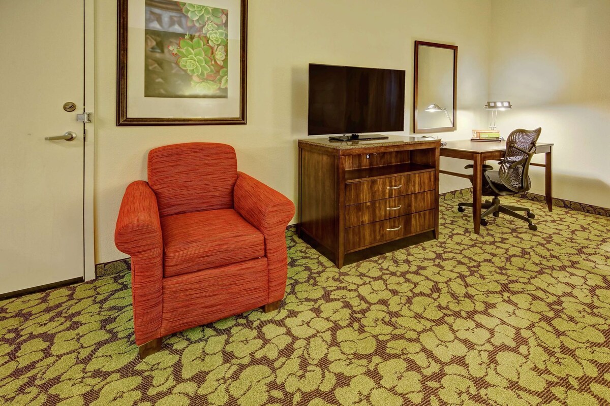 A hotel room with a red armchair, a wooden dresser, a desk with a chair, a television, and a mirror.