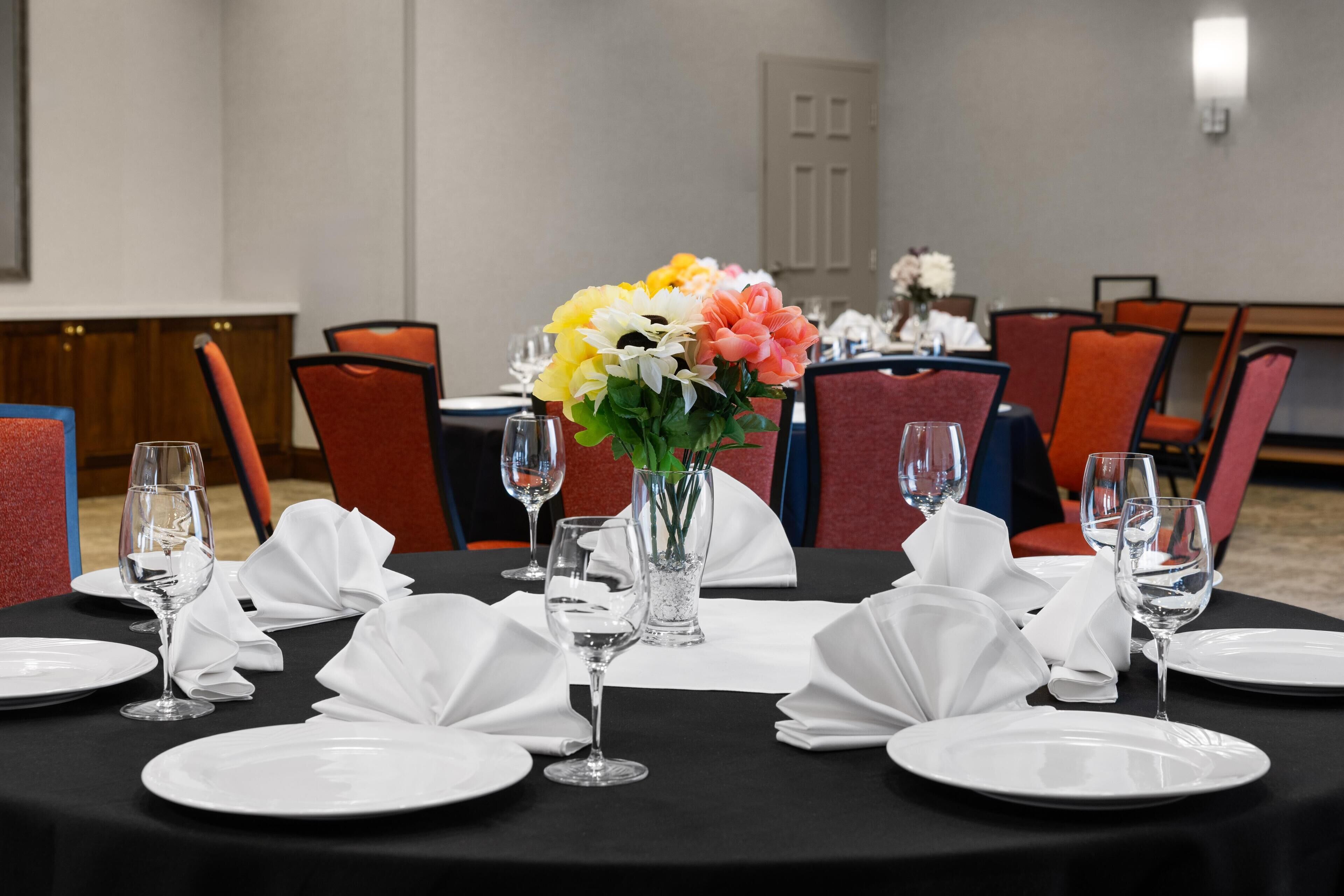 A conference room with round tables set for a meeting, equipped with white plates, glasses, and a centerpiece of colorful flowers.
