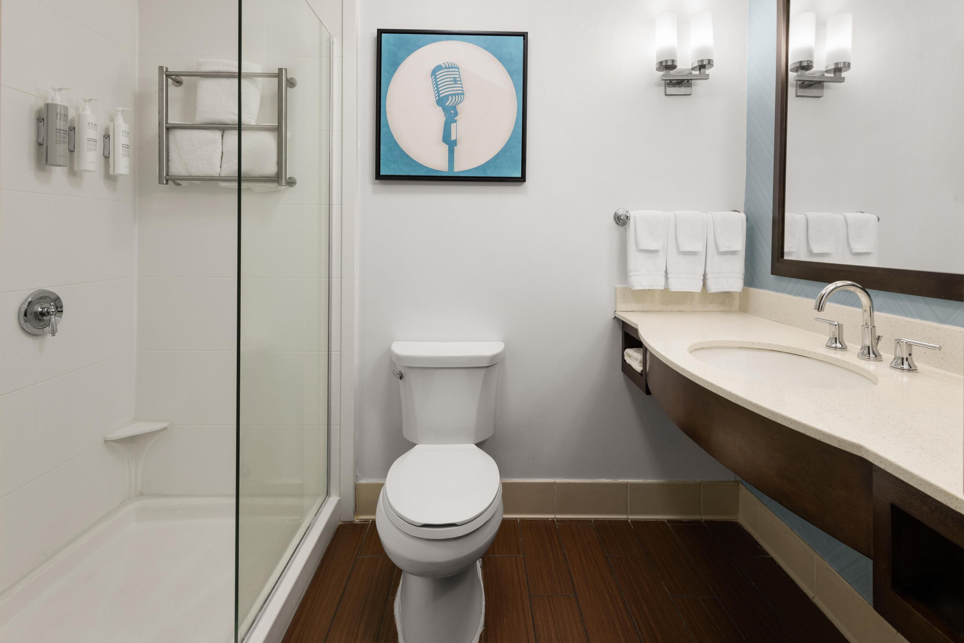 Une salle de bain moderne avec une douche à parois de verre, des toilettes blanches, un lavabo avec un miroir et une œuvre d’art encadrée accrochée au mur.