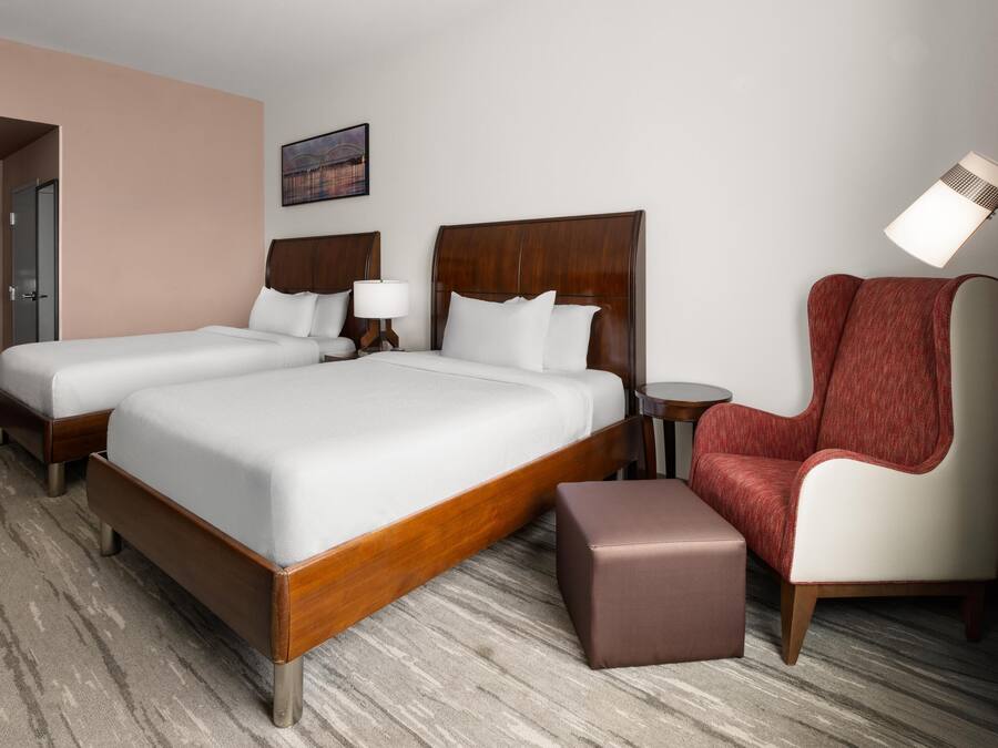 A hotel room with two beds, a red armchair, a small table, and a lamp.