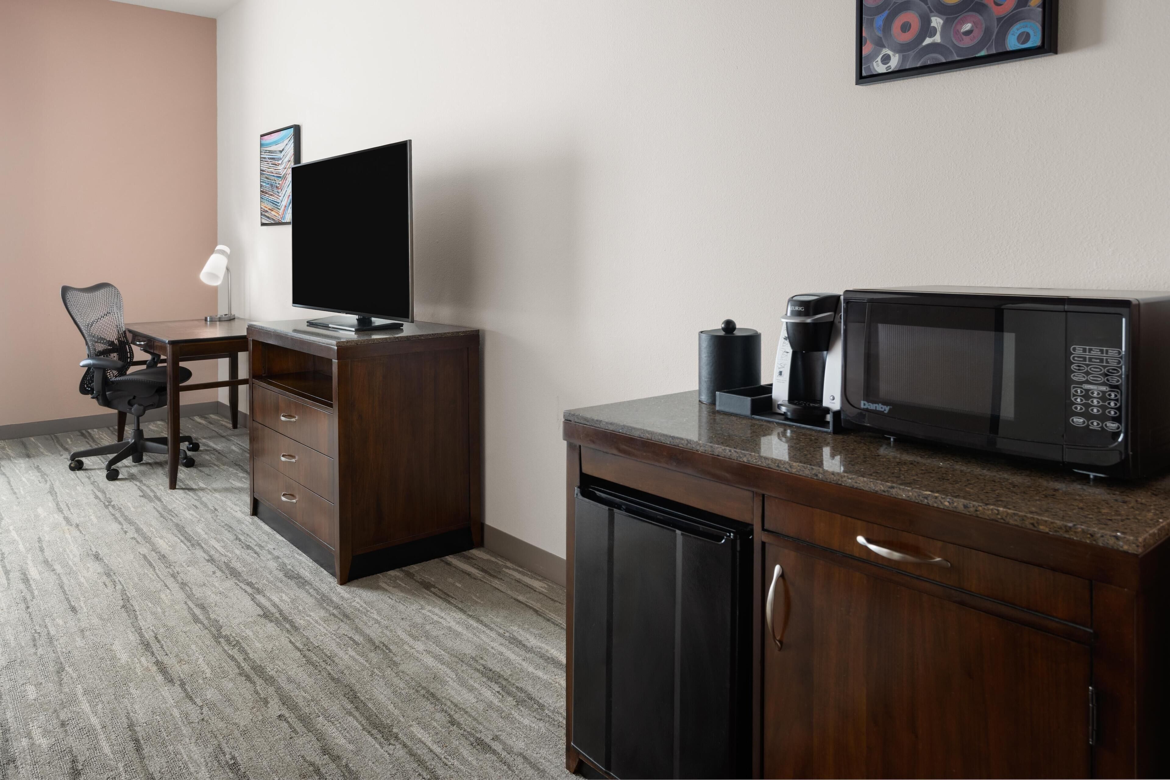 A hotel room with a wooden desk, a flat-screen TV, a microwave, a coffee maker, and a chair.