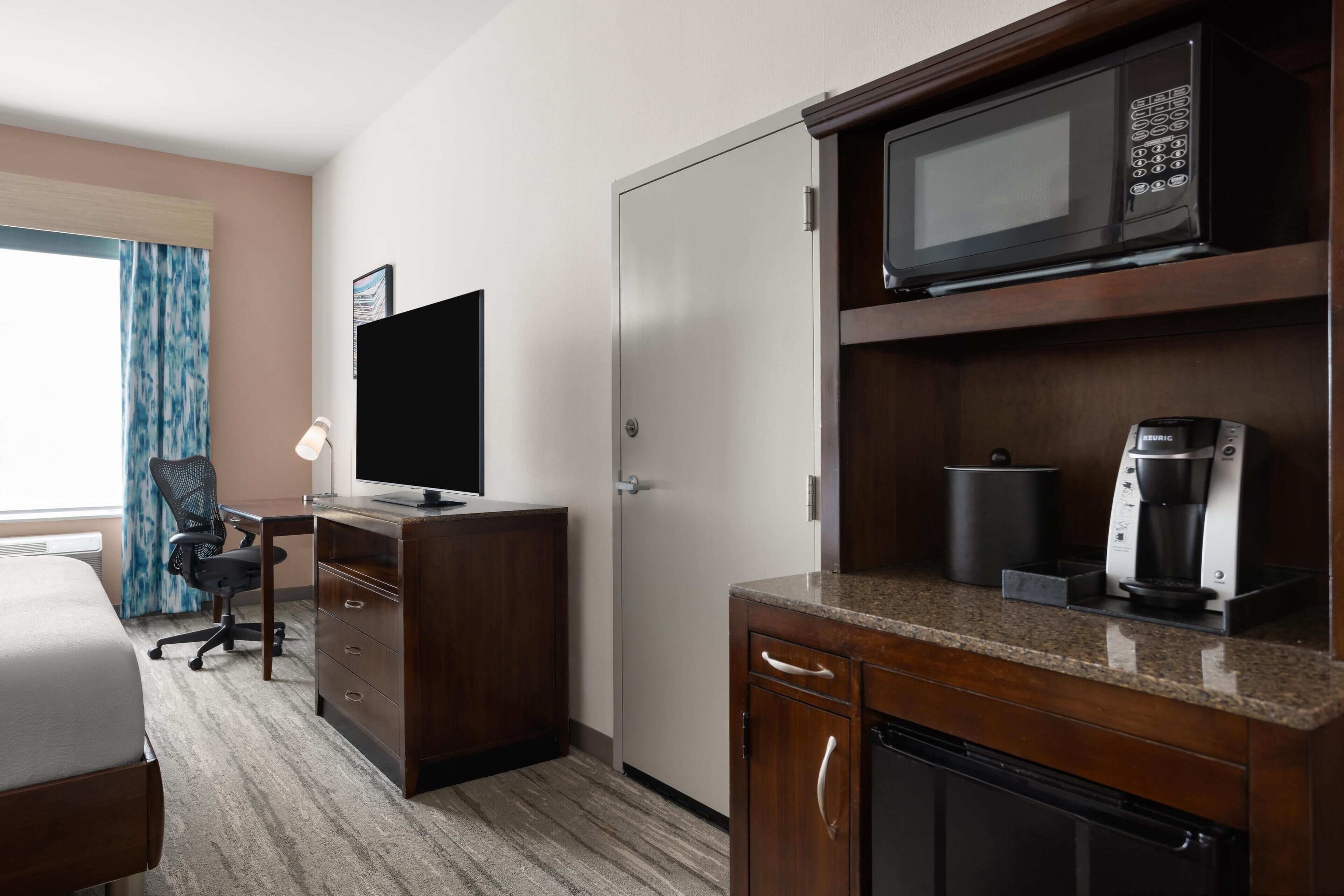Quarto de hotel com cama, escrivaninha, televisão, micro-ondas, máquina de café e guarda-roupa.