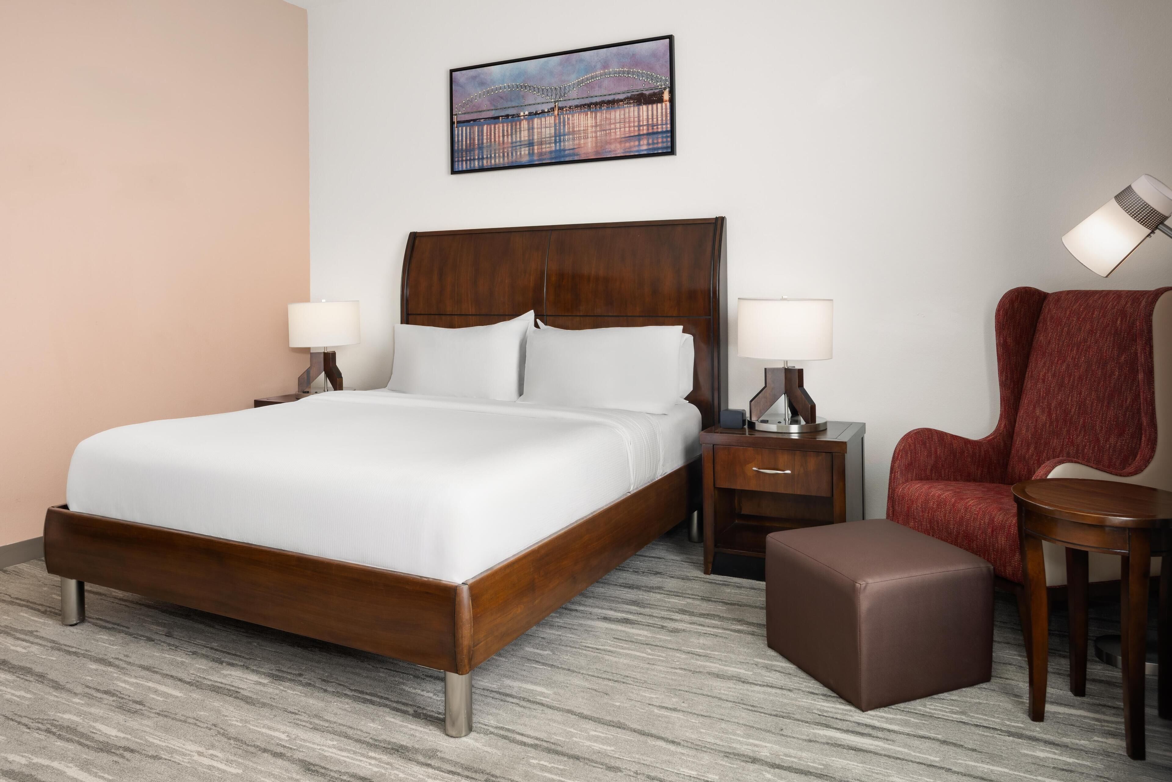 Habitación de hotel con una cama grande, dos mesitas de noche con lámparas, un sillón rojo, una pequeña mesa de madera y un cuadro en la pared.