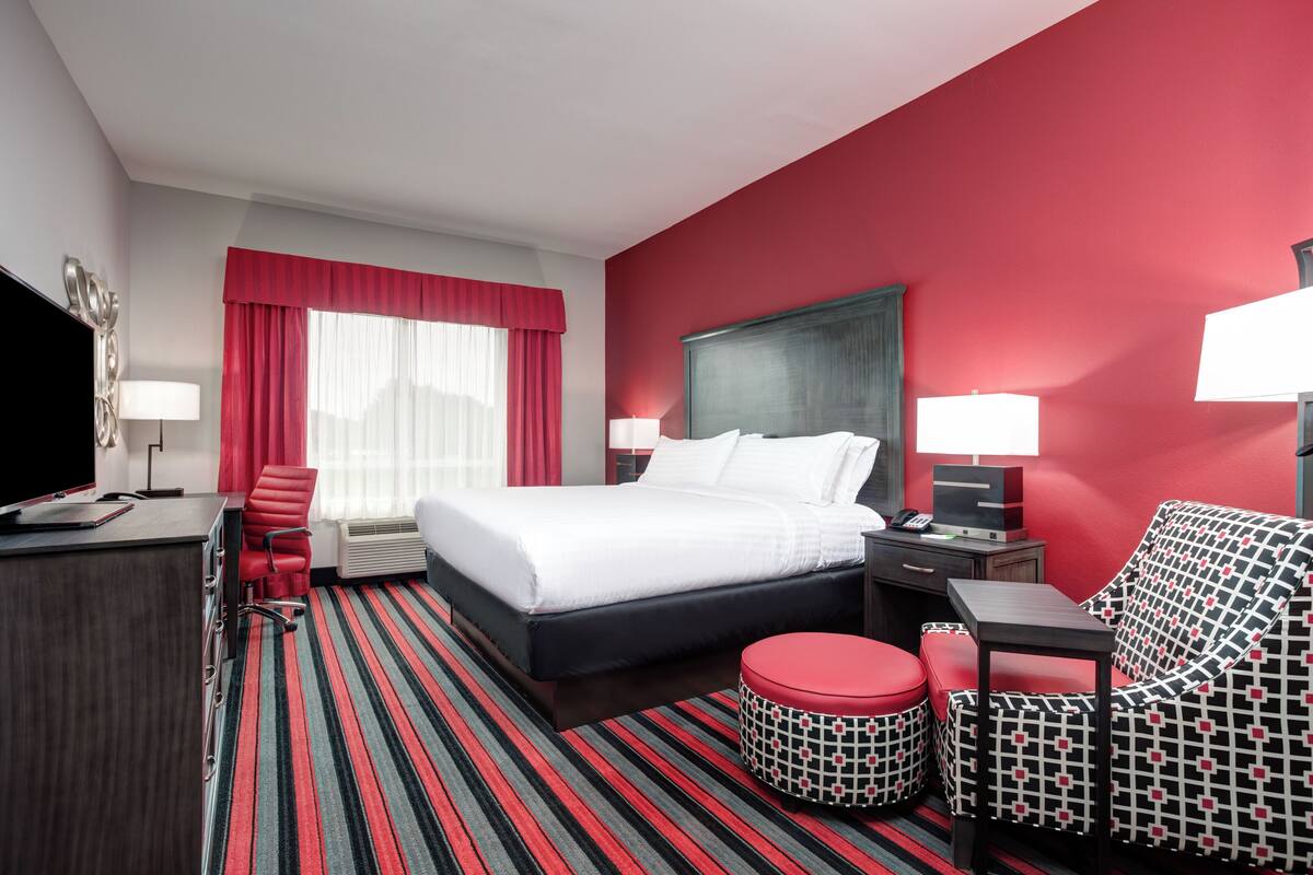 A hotel room with a large bed, a desk with a computer, a red chair, a patterned armchair, and a red ottoman.
