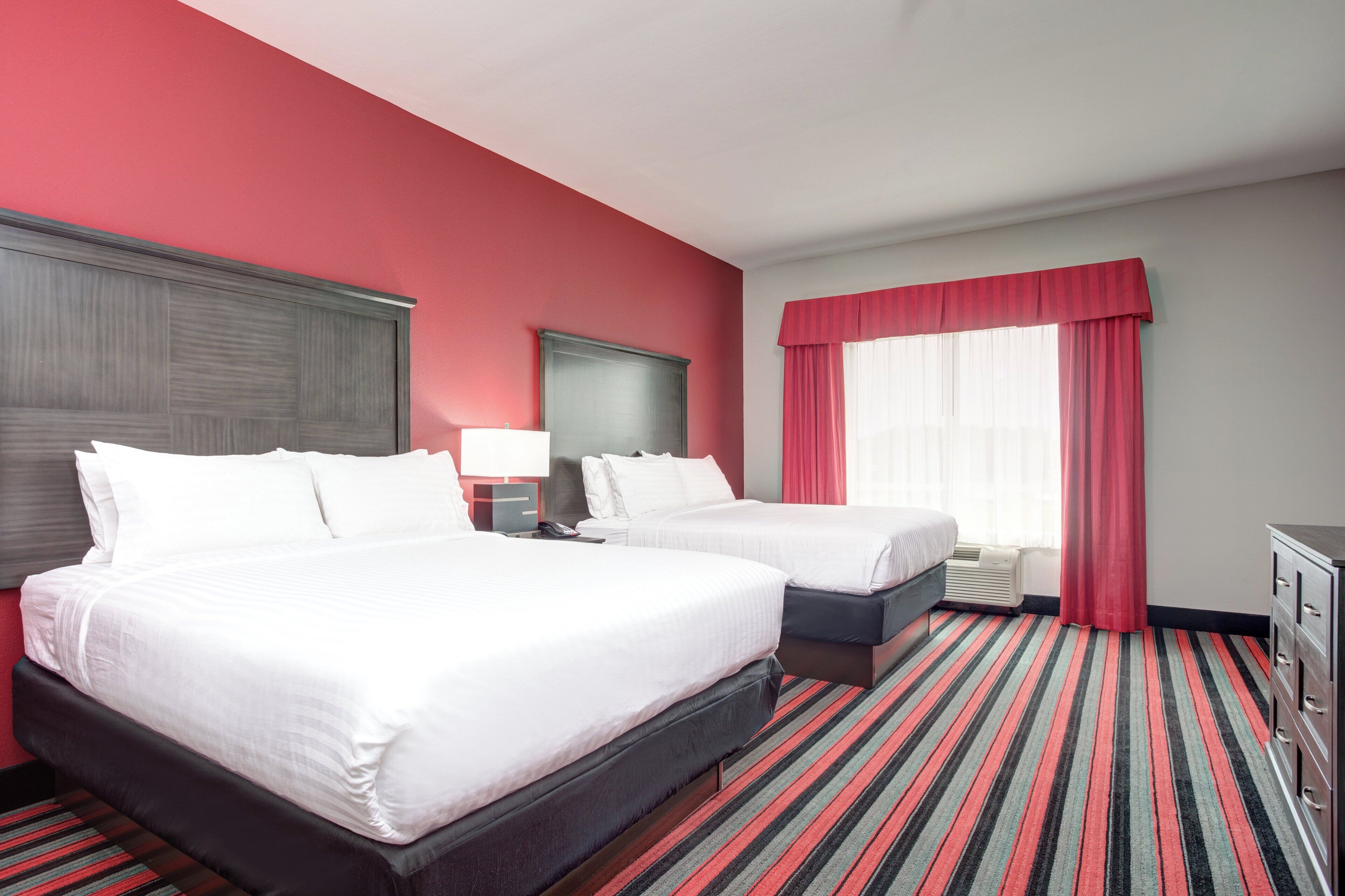 Habitación de hotel con dos camas, un escritorio, una silla, un ventanal con cortinas y una alfombra a rayas.