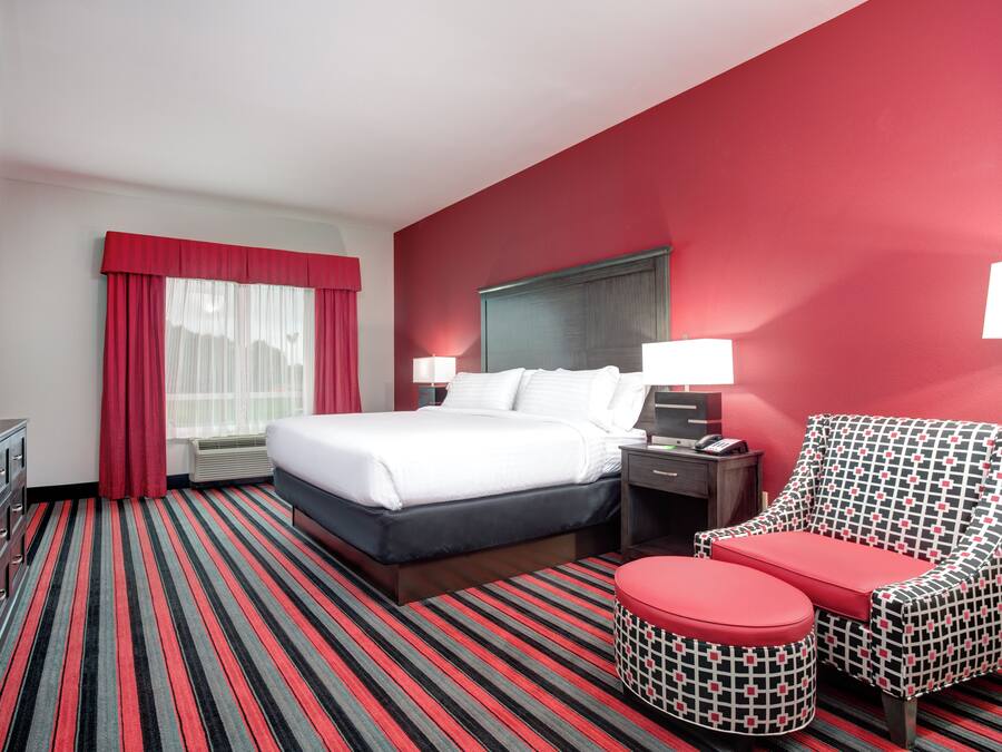 Habitación de hotel con cama, mesitas de noche, una silla con cojín rojo, alfombra a rayas y pared roja.