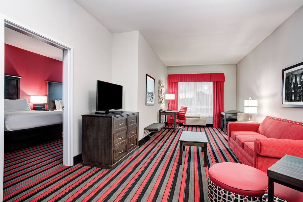 A hotel room with a bed, a TV, a red sofa, a dining table, and a red-striped carpet.