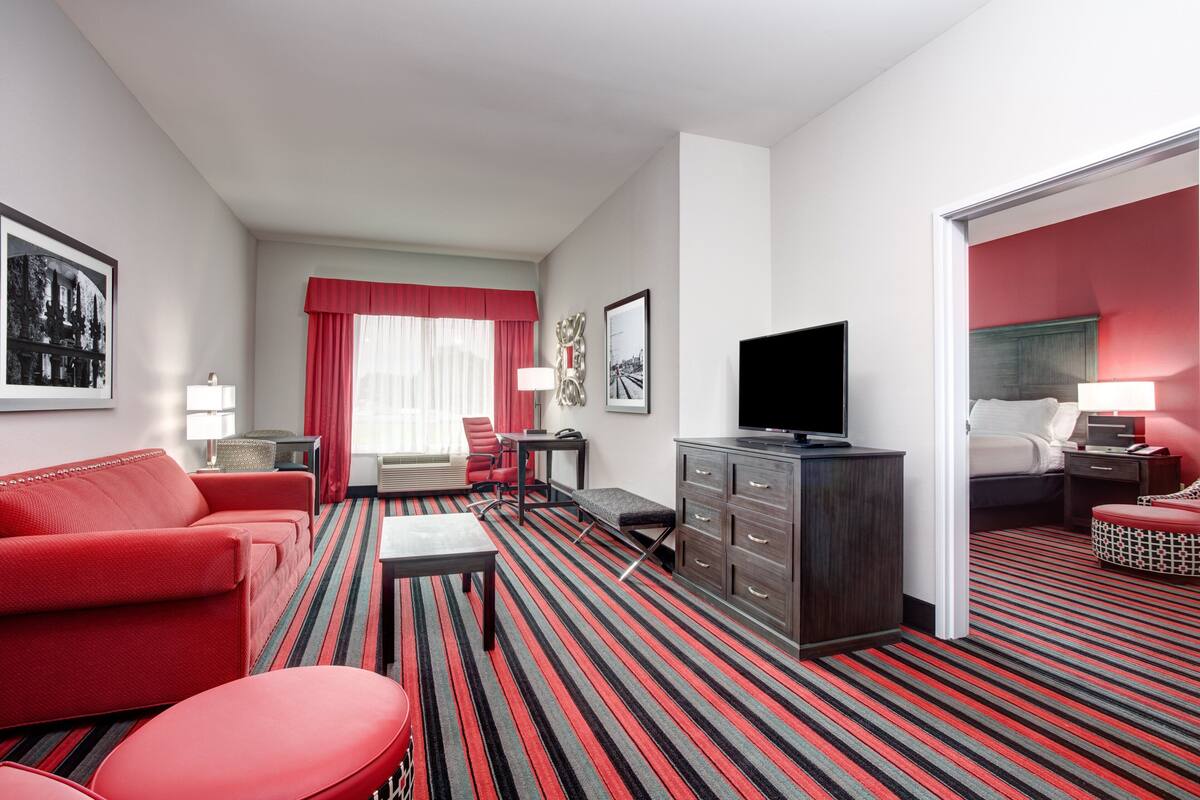 A hotel room with a red sofa, a TV, a desk with a lamp, and a bed with a red headboard.