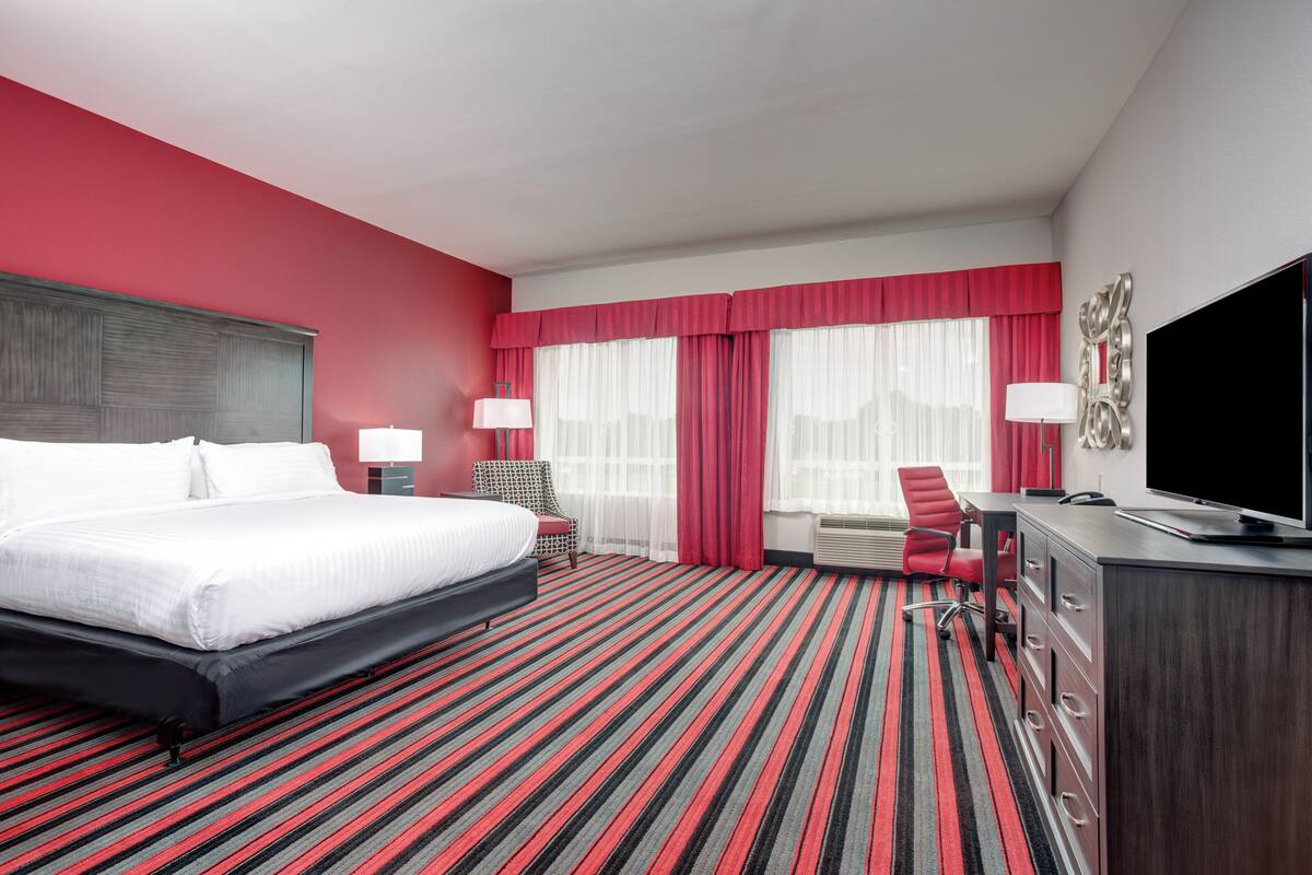 A hotel room with a large bed, a desk with a TV, a chair, and a red-striped carpet.